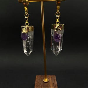 May include: Gold-coloured earrings with clear quartz crystal pendants, each with a small, purple amethyst accent. The earrings are displayed on a gold-coloured stand with a wooden base, against a dark background.