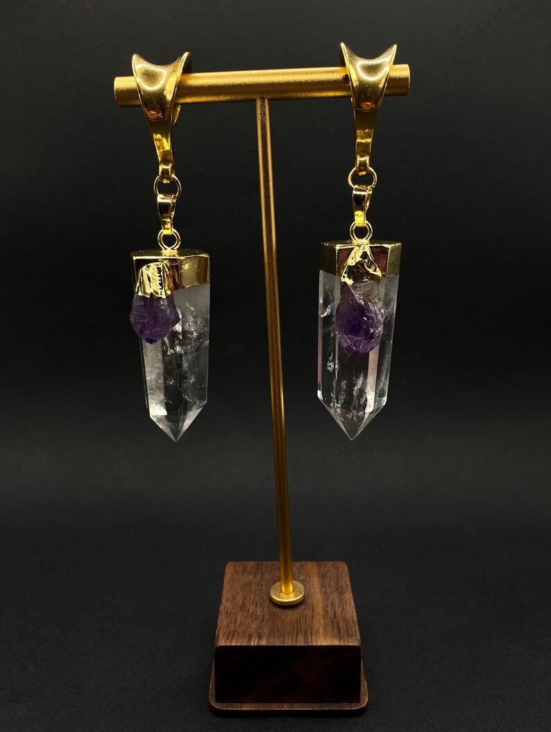 May include: Gold-coloured earrings with clear quartz crystal pendants, each with a small, purple amethyst accent. The earrings are displayed on a gold-coloured stand with a wooden base, against a dark background.