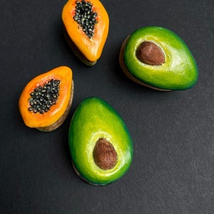 May include: Four hand-painted ceramic fruit sculptures, two green avocados and two orange papayas, on a black background.