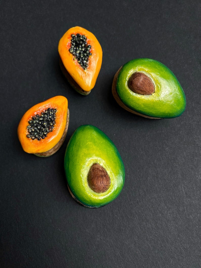 May include: Four hand-painted ceramic fruit sculptures, two green avocados and two orange papayas, on a black background.