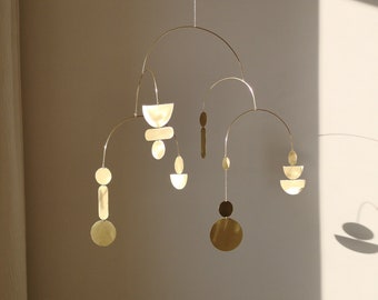 May include: A contemporary brass mobile featuring a series of geometric shapes, such as circles, semi-circles, and oblongs, suspended by fine wires. The mobile is displayed against a neutral wall, with its shadow visible. The shapes vary in size.