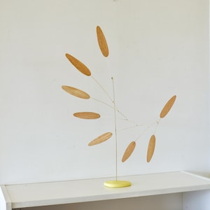 May include: A kinetic sculpture featuring a central vertical wire rod supported by a round yellow base. Seven thin, curved wire arms extend from the center, each holding a light brown, oval-shaped wooden leaf element in a balanced arrangement.