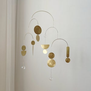 The photo shows a gold-colored brass kinetic mobile. the mobile is made of brass geometric shapes and transparent glass balls. the figures are connected using thin brass arches. mobile is suspended from the ceiling using a metal chain