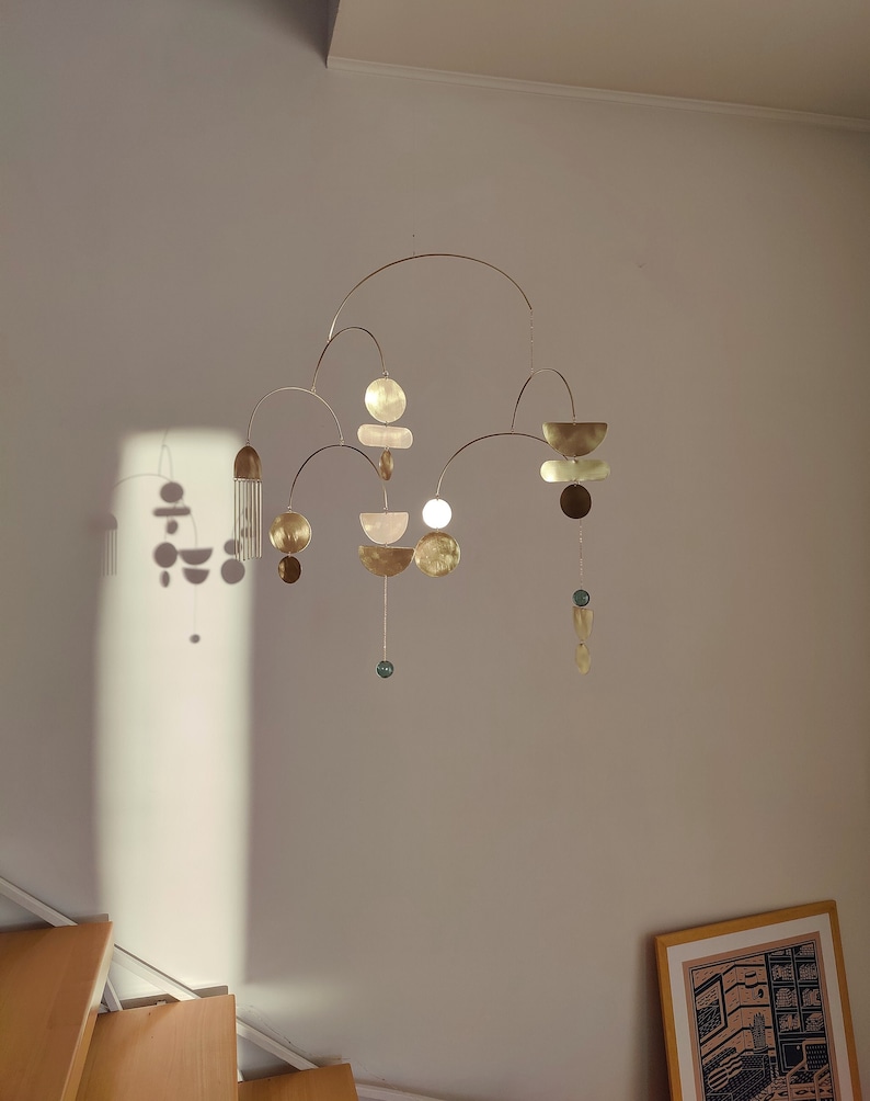 The photo shows a gold-colored brass kinetic mobile. the mobile is made of geometric circles, semicircles and ellipses of different shapes. the figures are connected using thin brass arches. mobile is suspended from the ceiling using a metal chain