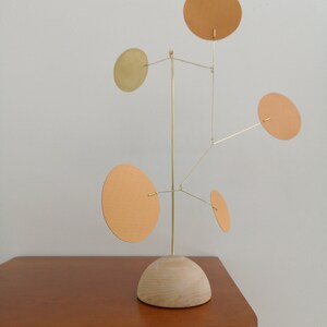 Op de afbeelding: Een kinetisch sculptuur met een centrale verticale messing staaf bevestigd op een halfronde basis van licht hout. Vijf dunne messing draden steken uit de staaf, elk met een platte, ronde schijf in verschillende tinten beige en goud.