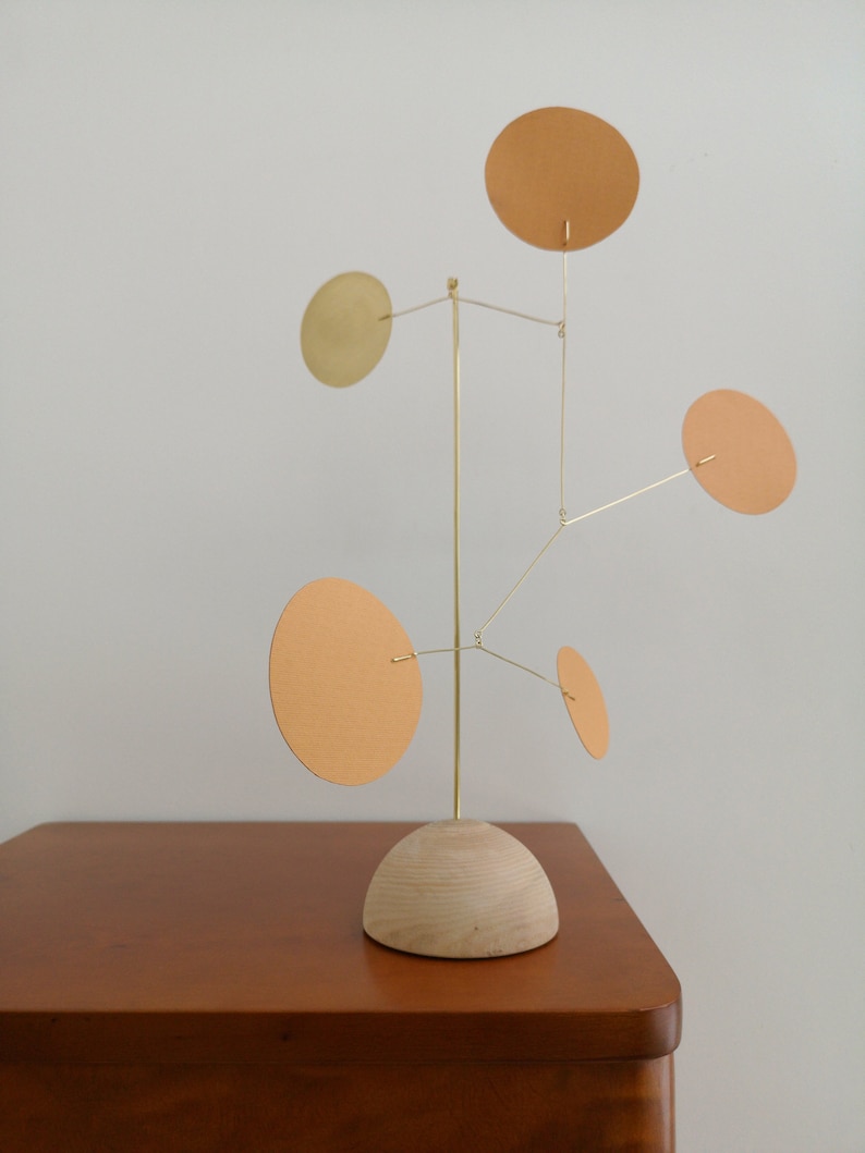 Op de afbeelding: Een kinetisch sculptuur met een centrale verticale messing staaf bevestigd op een halfronde basis van licht hout. Vijf dunne messing draden steken uit de staaf, elk met een platte, ronde schijf in verschillende tinten beige en goud.