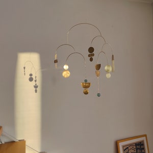 The photo shows a gold-colored brass kinetic mobile. the mobile is made of geometric circles, semicircles and ellipses of different shapes. the figures are connected using thin brass arches. mobile is suspended from the ceiling using a metal chain