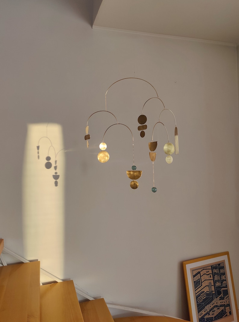 The photo shows a gold-colored brass kinetic mobile. the mobile is made of geometric circles, semicircles and ellipses of different shapes. the figures are connected using thin brass arches. mobile is suspended from the ceiling using a metal chain