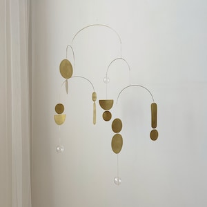 The photo shows a gold-colored brass kinetic mobile. the mobile is made of geometric circles, semicircles and ellipses of different shapes. the figures are connected using thin brass arches. mobile is suspended from the ceiling using a metal chain