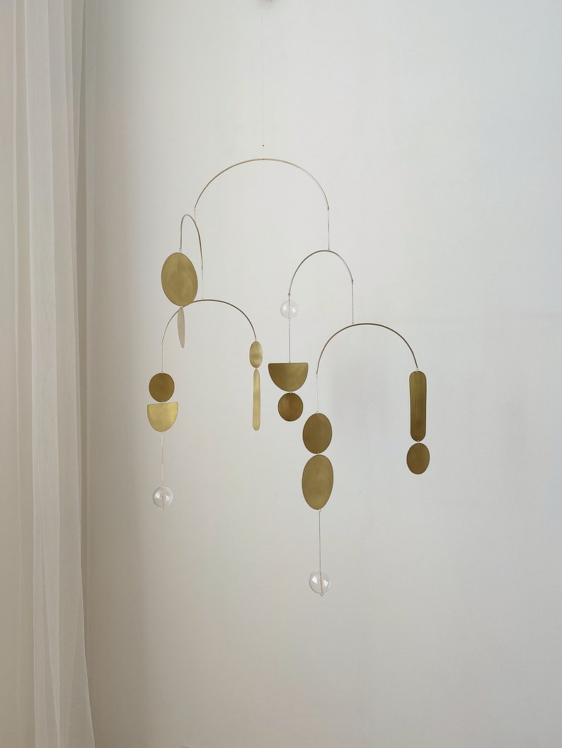 The photo shows a gold-colored brass kinetic mobile. the mobile is made of geometric circles, semicircles and ellipses of different shapes. the figures are connected using thin brass arches. mobile is suspended from the ceiling using a metal chain