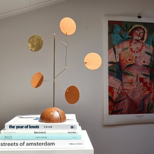 Op de afbeelding: Een tafel-mobielsculptuur met een halfronde houten voet en een verticale messing stang. Vijf dunne messing armen steken uit de stang, elk voorzien van een platte ronde schijf in tinten van beige, oranje en metallic goud, verticaal gebalanceerd.