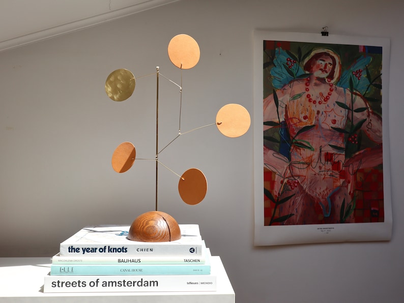 Op de afbeelding: Een tafel-mobielsculptuur met een halfronde houten voet en een verticale messing stang. Vijf dunne messing armen steken uit de stang, elk voorzien van een platte ronde schijf in tinten van beige, oranje en metallic goud, verticaal gebalanceerd.