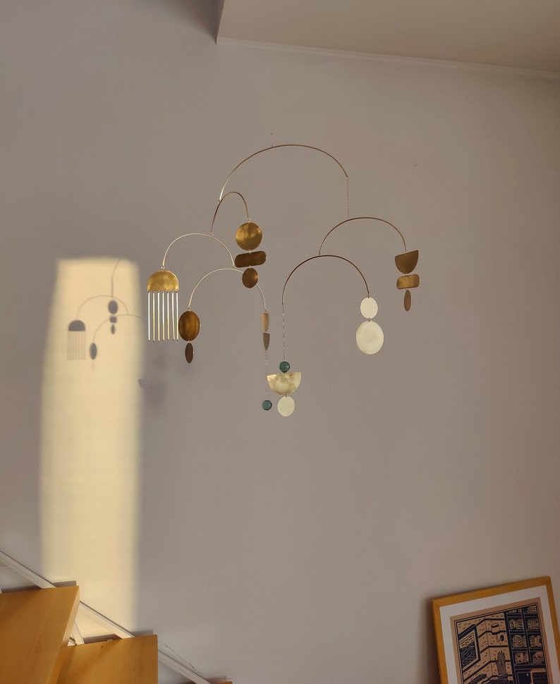 The photo shows a gold-colored brass kinetic mobile. the mobile is made of geometric circles, semicircles and ellipses of different shapes. the figures are connected using thin brass arches. mobile is suspended from the ceiling using a metal chain