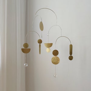 The photo shows a gold-colored brass kinetic mobile. the mobile is made of geometric circles, semicircles and ellipses of different shapes. the figures are connected using thin brass arches. mobile is suspended from the ceiling using a metal chain