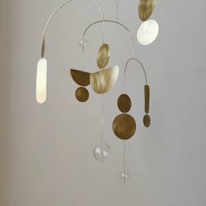 The photo shows a gold-colored brass kinetic mobile. the mobile is made of geometric circles, semicircles and ellipses of different shapes. the figures are connected using thin brass arches. mobile is suspended from the ceiling using a metal chain