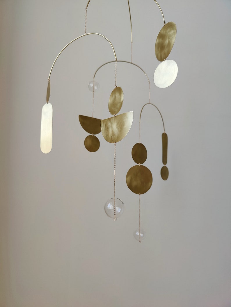 The photo shows a gold-colored brass kinetic mobile. the mobile is made of geometric circles, semicircles and ellipses of different shapes. the figures are connected using thin brass arches. mobile is suspended from the ceiling using a metal chain