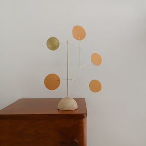 Op de afbeelding: Een tafelmobielsculptuur met een lichtgekleurde houten koepelvormige voet en een dun frame van messing draad. Vijf ronde schijven zijn bevestigd aan de draadarmen, met vier schijven in een matte oranje kleur en &eacute;&eacute;n schijf in een metallic goud.