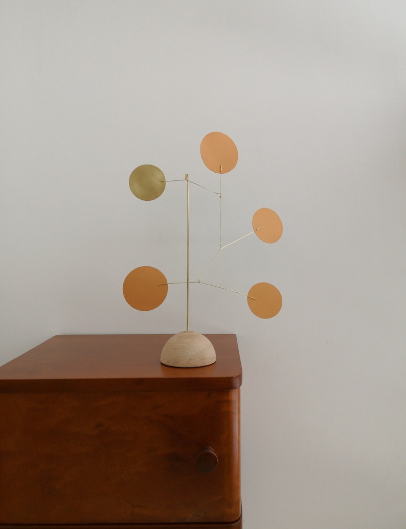 Op de afbeelding: Een tafelmobielsculptuur met een lichtgekleurde houten koepelvormige voet en een dun frame van messing draad. Vijf ronde schijven zijn bevestigd aan de draadarmen, met vier schijven in een matte oranje kleur en &eacute;&eacute;n schijf in een metallic goud.