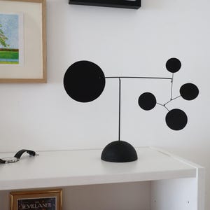 May include: A black metal mobile sculpture featuring a large disc and smaller circles connected by thin rods. The sculpture rests on a rounded base, offering a modern, minimalist design. The artwork is displayed on a white shelf, in cm.