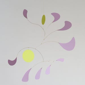 May include: A hanging mobile featuring thin metal wires connected to several flat, lavender-colored petal shapes and two solid circular discs, one in a bright lime green and the other in a lighter yellow-green, suspended in a balanced arrangement.