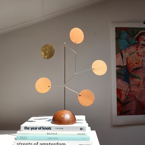 Op de afbeelding: Een tafelmobiel-sculptuur met een halfronde voet van donker hout, een dunne verticale metalen staaf en vijf hangende ronde schijven. Vier schijven zijn mat beige van kleur, terwijl &eacute;&eacute;n schijf is gemaakt van reflecterend goudkleurig metaal.