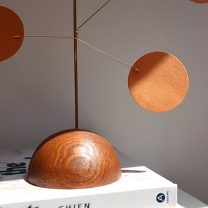 Op de afbeelding: Een tafelmobiel met een donkerhouten halfronde voet en een structuur van dun messingdraad. Drie oranje cirkelvormige schijven met een getextureerd oppervlak hangen aan de draadarmen, wat zorgt voor een gebalanceerde compositie van vormen.