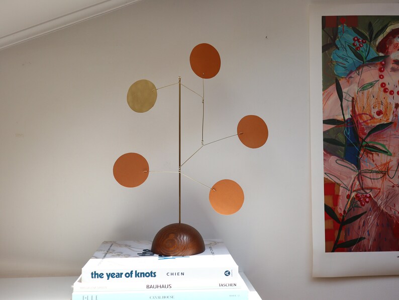 Op de afbeelding: Een mobiele sculptuur voor op tafel met een halfronde voet van donker hout en een dunne verticale messing stang. Vijf dunne, ronde schijven in terracotta oranje en gedempt goud hangen aan fijne messing draden aan de centrale stang.