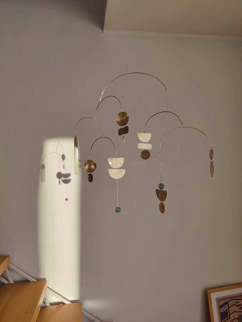 The photo shows a gold-colored brass kinetic mobile. the mobile is made of geometric circles, semicircles and ellipses of different shapes. the figures are connected using thin brass arches. mobile is suspended from the ceiling using a metal chain