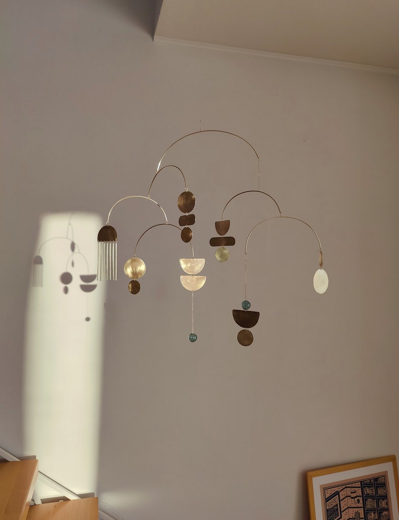 The photo shows a gold-colored brass kinetic mobile. the mobile is made of geometric circles, semicircles and ellipses of different shapes. the figures are connected using thin brass arches. mobile is suspended from the ceiling using a metal chain