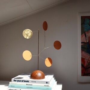 Op de afbeelding: Een tafelmodel mobiel sculptuur met een donkerhouten halfronde voet, een dunne verticale metalen staaf en vijf dunne metalen armen. Elke arm draagt een ronde schijf in verschillende tinten messing, oranje en lichtbruin.