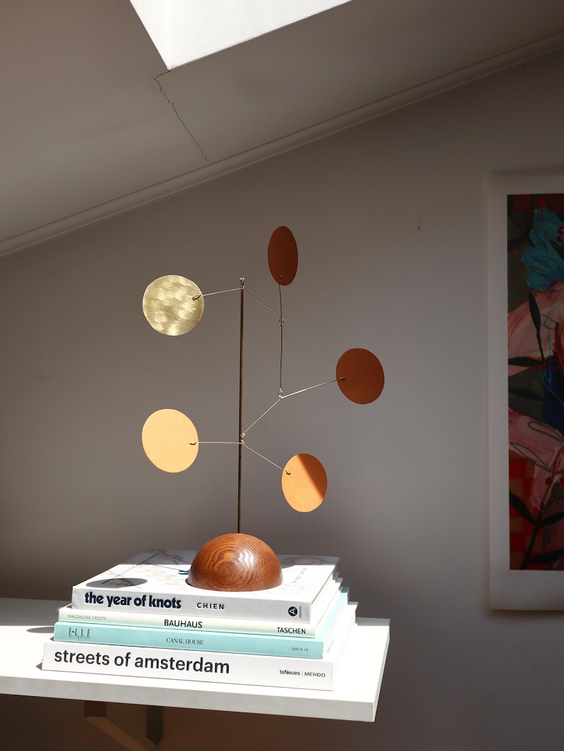 Op de afbeelding: Een tafelmodel mobiel sculptuur met een donkerhouten halfronde voet, een dunne verticale metalen staaf en vijf dunne metalen armen. Elke arm draagt een ronde schijf in verschillende tinten messing, oranje en lichtbruin.