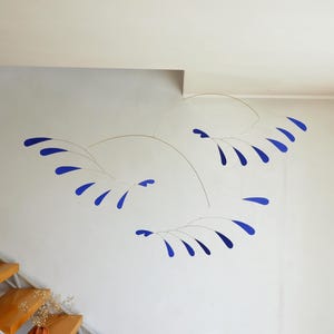May include: A hanging mobile sculpture featuring thin, curved metal wires that support multiple small, flat, blue, teardrop-shaped elements arranged in three distinct, sweeping, branch-like clusters that extend across the wall space.