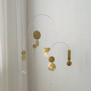 The photo shows a gold-colored brass kinetic mobile. the mobile is made of brass geometric shapes and transparent glass balls. the figures are connected using thin brass arches. mobile is suspended from the ceiling using a metal chain