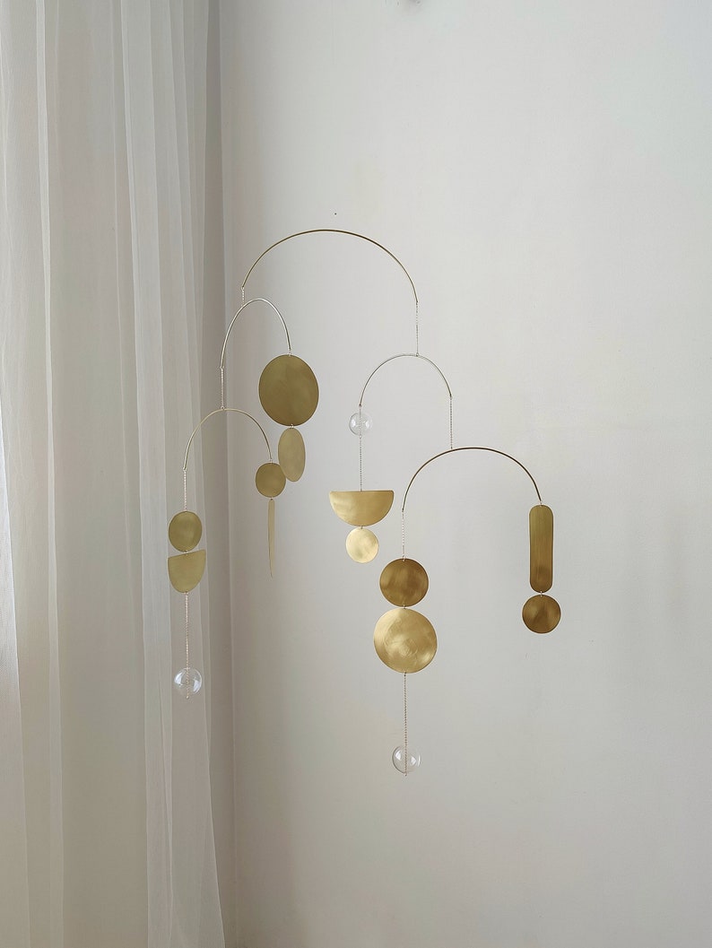 The photo shows a gold-colored brass kinetic mobile. the mobile is made of brass geometric shapes and transparent glass balls. the figures are connected using thin brass arches. mobile is suspended from the ceiling using a metal chain