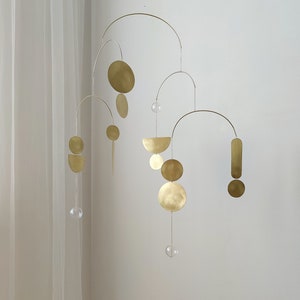 The photo shows a gold-colored brass kinetic mobile. the mobile is made of geometric circles, semicircles and ellipses of different shapes. the figures are connected using thin brass arches. mobile is suspended from the ceiling using a metal chain