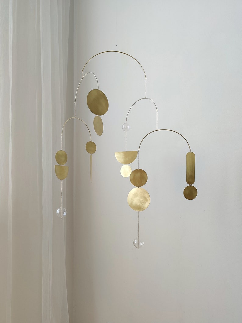 The photo shows a gold-colored brass kinetic mobile. the mobile is made of geometric circles, semicircles and ellipses of different shapes. the figures are connected using thin brass arches. mobile is suspended from the ceiling using a metal chain