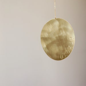 Op de afbeelding: Een cirkelvormige goudkleurige metalen hanger met een geborstelde afwerking, hangend aan een dunne bijpassende ketting. Het oppervlak van de schijf bevat de gegraveerde datum 03.07.22 langs de rechterrand, met een metalen textuur en glans.