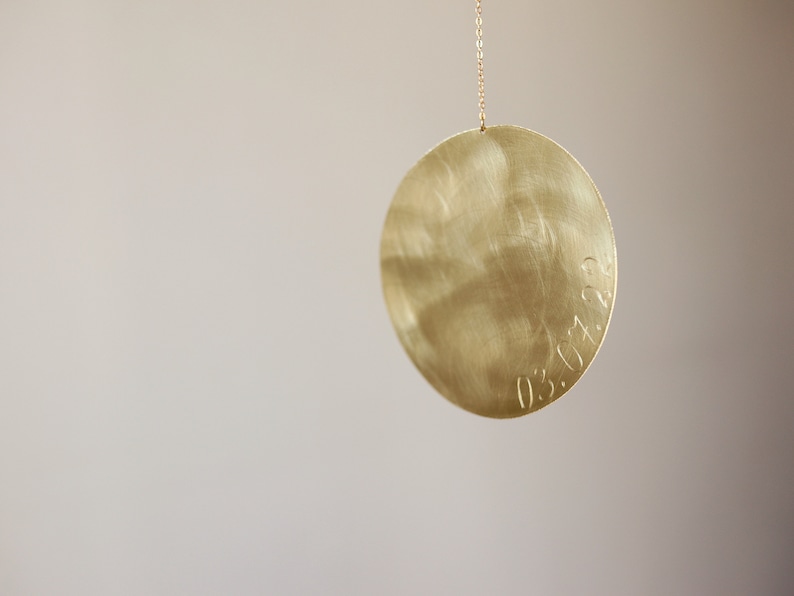Op de afbeelding: Een cirkelvormige goudkleurige metalen hanger met een geborstelde afwerking, hangend aan een dunne bijpassende ketting. Het oppervlak van de schijf bevat de gegraveerde datum 03.07.22 langs de rechterrand, met een metalen textuur en glans.