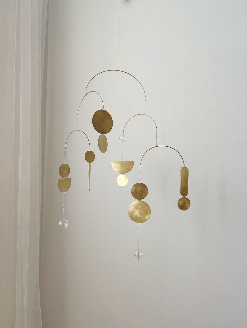 The photo shows a gold-colored brass kinetic mobile. the mobile is made of brass geometric shapes and transparent glass balls. the figures are connected using thin brass arches. mobile is suspended from the ceiling using a metal chain