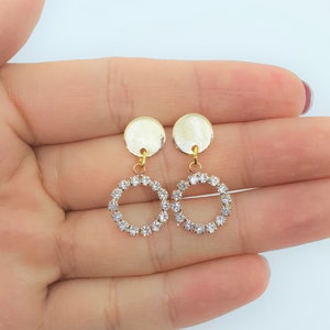 May include: A pair of gold earrings with a round, clear rhinestone design. The earrings have a small, round, gold disc at the top.