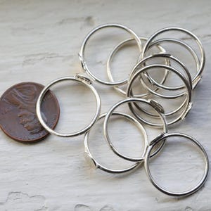 May include: A collection of silver-colored rings, some overlapping, are displayed next to a copper-colored penny. The rings are thin and circular, with a simple, minimalist design. The penny provides a size reference.