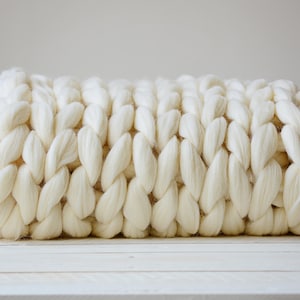 May include: A white chunky knit throw blanket made of thick yarn. The blanket is rolled up and sitting on a white wooden surface.