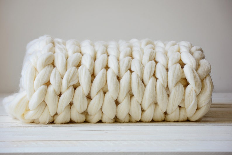 May include: A white chunky knit throw blanket made of thick yarn. The blanket is rolled up and sitting on a white wooden surface.