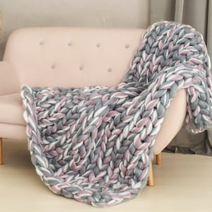 May include: A chunky knit throw blanket in shades of gray, white, and pink, draped over a light pink sofa.