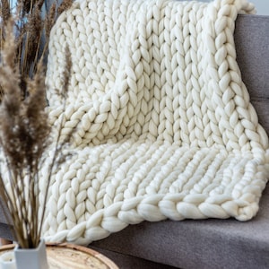 K&ouml;nnte beinhalten: Eine wei&szlig;e, grob gestrickte Wolldecke, die &uuml;ber ein graues Sofa gelegt ist. Die Decke ist aus dickem Garn gefertigt und hat eine weiche, kuschelige Textur.