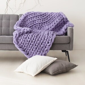 May include: A large, chunky knit throw blanket in a light purple color draped over a gray sofa. The blanket is made of thick yarn and has a soft, cozy texture.