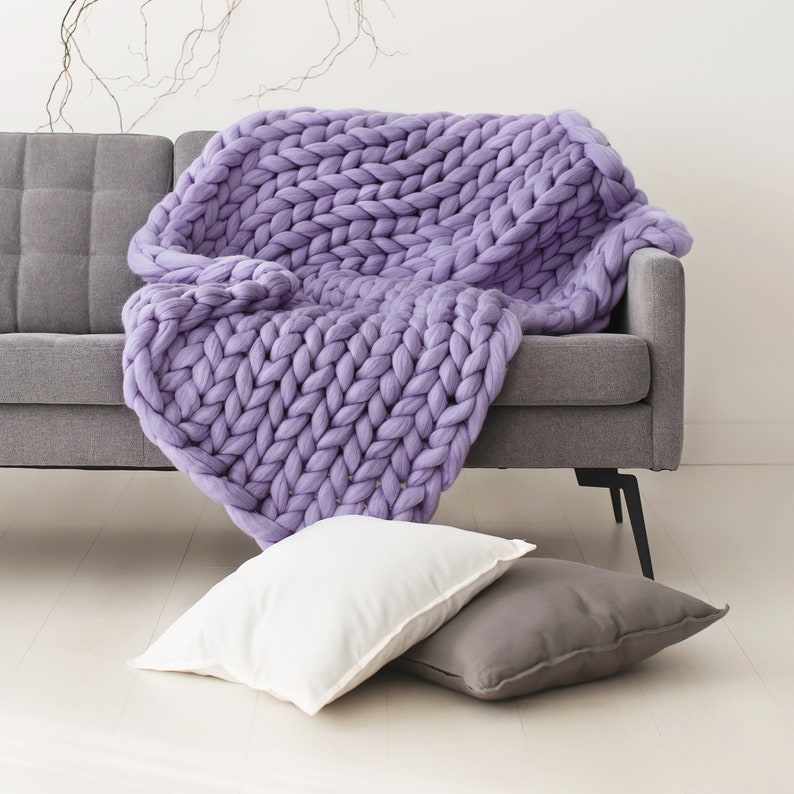 May include: A large, chunky knit throw blanket in a light purple color draped over a gray sofa. The blanket is made of thick yarn and has a soft, cozy texture.
