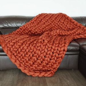 May include: A chunky knit throw blanket in a burnt orange colour. The blanket is made of thick yarn and has a soft, cosy texture.