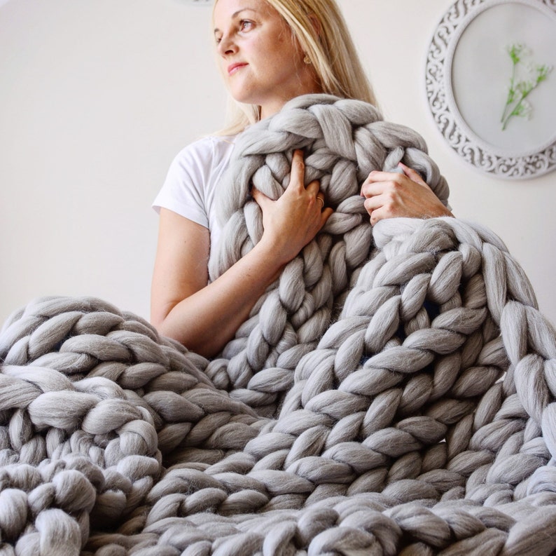 May include: A large, chunky knit blanket in a light gray color. The blanket is made of thick yarn and has a soft, cozy texture.