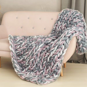 K&ouml;nnte beinhalten: Eine dicke Strickdecke in den Farben Grau, Wei&szlig; und Rosa, &uuml;ber ein hellrosa Sofa gelegt.